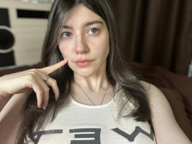 VinnaSkyHott with long brown hair, light skin, and green eyes, pouting lips, wearing a white tank top, touching cheek with finger, showing a ring. Boobs visible, brown curtains.