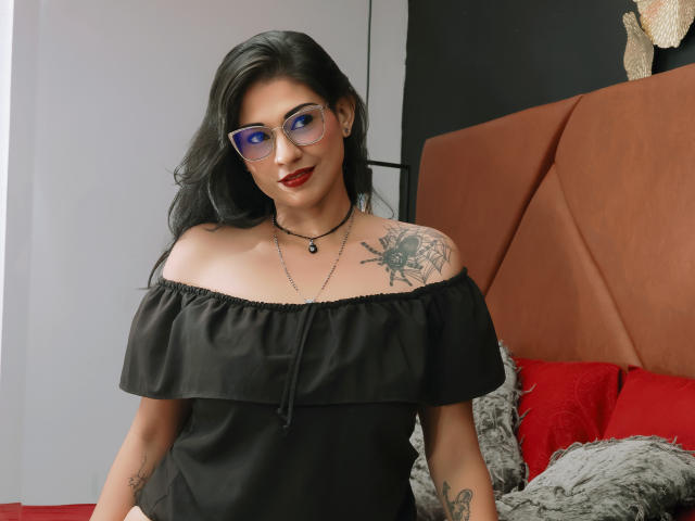 IvanaVelvet with black off-shoulder top, glasses, red lipstick, tattoos on shoulder and arm, necklace, smiling, standing in bedroom with red pillows and gray headboard.