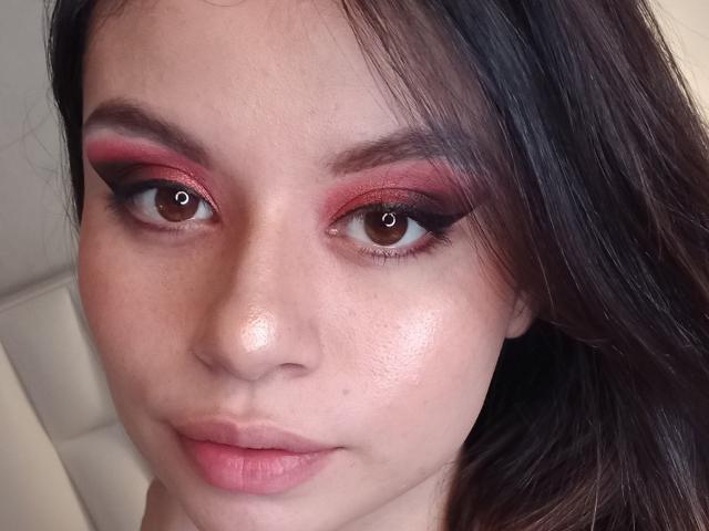 KennyAndLucy has glossy brown eyes with red and pink eyeshadow, full pink lips, and shiny skin, with long dark brown hair on the right side of the face.