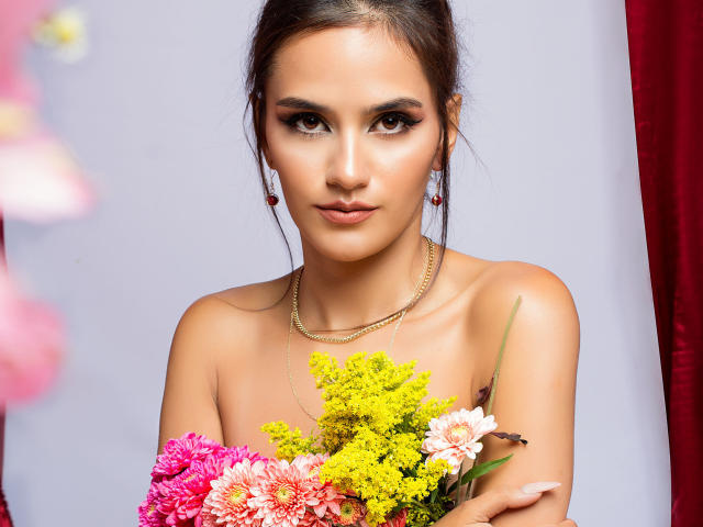 AmelieLautrec with tan skin, brown eyes, and dark hair, holding colorful flowers, wearing gold jewelry, and looking directly at the camera, with pink and yellow flowers in front.