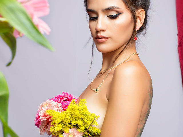 AmelieLautrec with pouty lips, dark eyeliner, and pink lipstick, holding a bouquet of pink, yellow, and green flowers, wearing a gold necklace and red earrings, topless.
