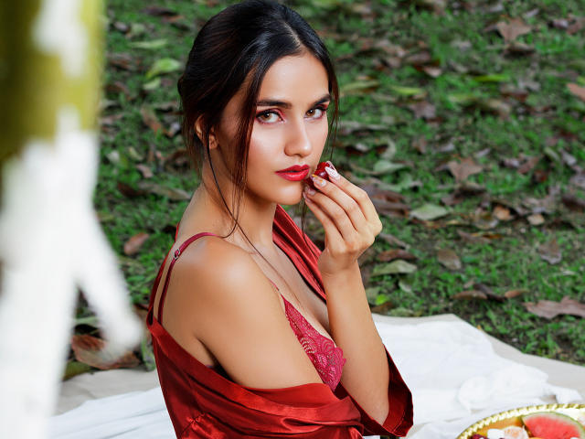 AmelieLautrec with dark hair, red lipstick, and red lace bra, holding a red apple to her mouth, wearing a red silk robe, sitting on a white blanket.