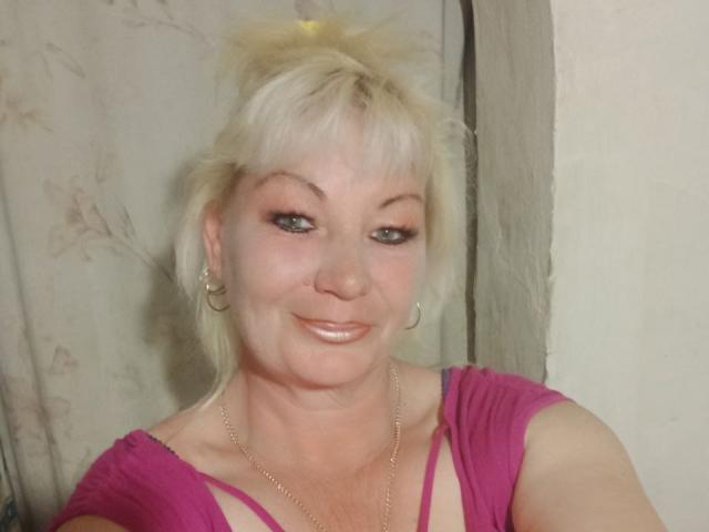 KathyNettie with blonde hair, green eyes, and a pink top, showing her tits in a selfie, smiling with a nose ring and hoop earrings, wearing a gold chain.