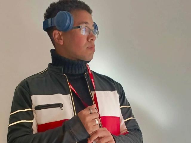 MartinxHt has dark skin, curly black hair, wears blue headphones, glasses, and a black turtleneck with a zip-up jacket featuring red, white, and black stripes.
