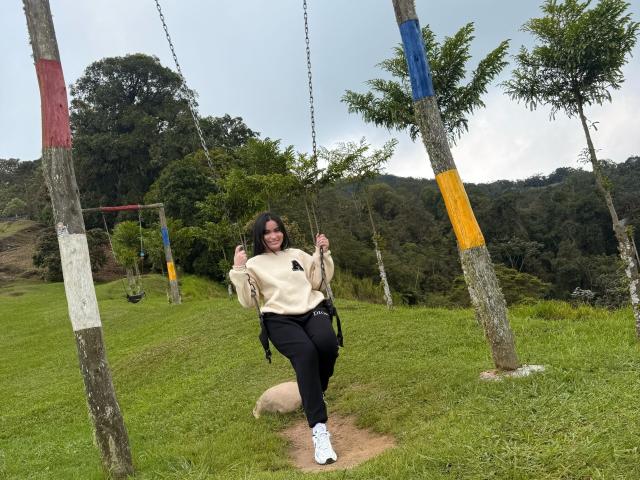 Miia Palmer, con i capelli scuri e un sorriso, è in piedi su un sentiero sterrato, tenendo le catene di un'altalena, indossa un maglione beige, pantaloni neri e scarpe da ginnastica bianche, in un parco erboso..