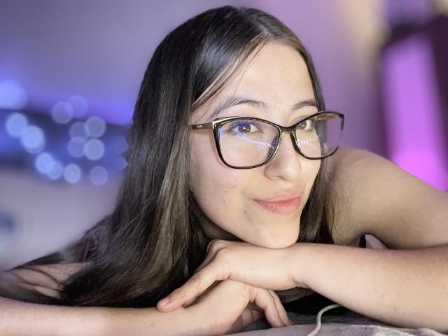 KatiaRogers with long dark hair, wearing glasses, resting her chin on her hands, smiling softly, showing small moles on her cheek, in a well-lit, slightly blurred room.