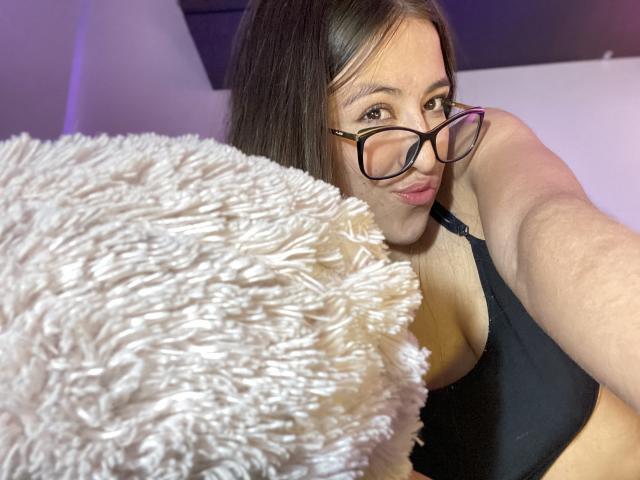 KatiaRogers with glasses, black bra, and fluffy white pillow, taking a selfie. Her tits are visible in the black bra, and she's looking at the camera.