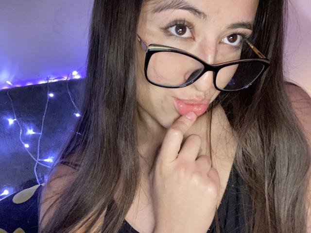 KatiaRogers with long brown hair, wearing black glasses, pouting with finger on her pink, glossy lips, looking at the camera. Her ass is slightly visible.