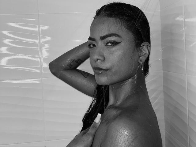 Black-and-white photo of ChloeBrownOne with wet, slicked-back hair, looking at the camera, water droplets on her skin, hand behind head, earrings, and a tattoo on her upper arm.