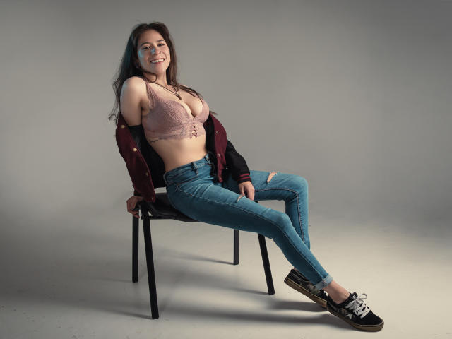 EveeDreams with big tits, wearing a pink lace bra, ripped jeans, and black sneakers, sitting on a chair, smiling, with a maroon jacket off her shoulders.