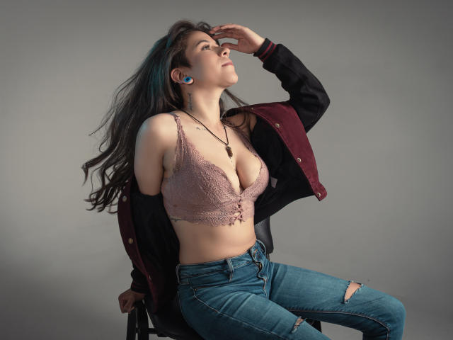 EveeDreams with long dark hair, wearing a pink lace bra, black and maroon jacket, and ripped jeans, sits on a chair, hand over her eyes, showing her tits.
