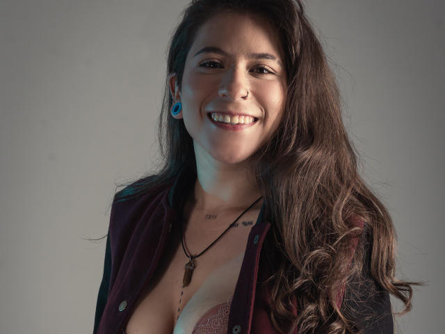 EveeDreams, smiling with long brown hair, blue ear gauges, maroon shirt, necklace, visible cleavage, and a tattoo on her chest. Her tits are partially shown.