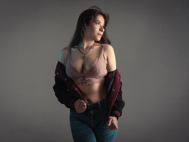 EveeDreams with big boobs in a pink lace bra, unbuttoned maroon jacket, and blue jeans, looking to the side, has tattoos and blue earbuds, standing in a gray room.