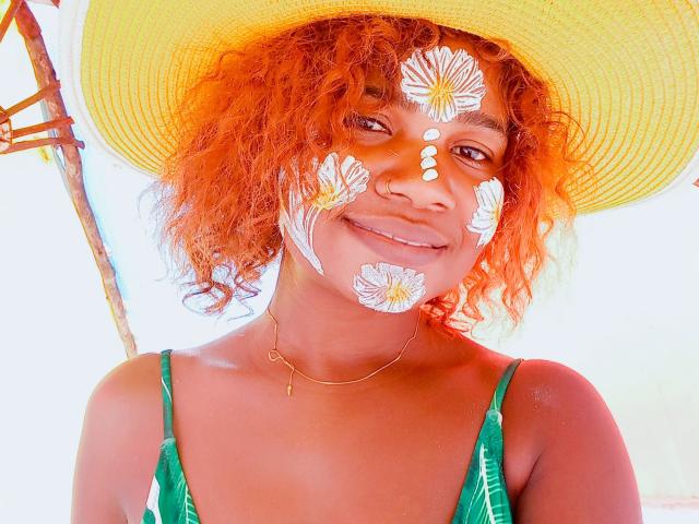 Verocka with orange curly hair, wearing a green dress, yellow hat, and white flower face paint, smiling. Her tits are covered by the dress, face paint on her nose.