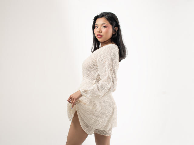 LizzyClay vestida con un vestido blanco transparente de punto, mostrando una vista lateral de sus pequeños pechos y su trasero desnudo, con cabello negro y lápiz labial rojo, de pie frente a un fondo blanco..