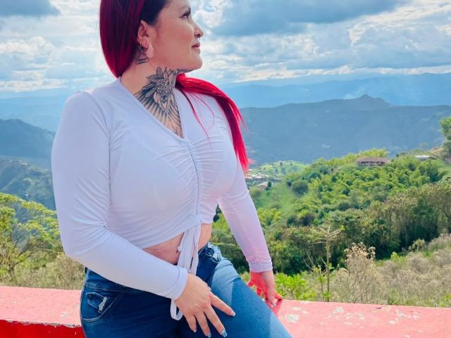 Isabella Dankworth ha i capelli rossi, indossa un top bianco e jeans blu, mostrando un grande tatuaggio sul collo. Il suo seno è visibile, sullo sfondo di una vista montuosa..