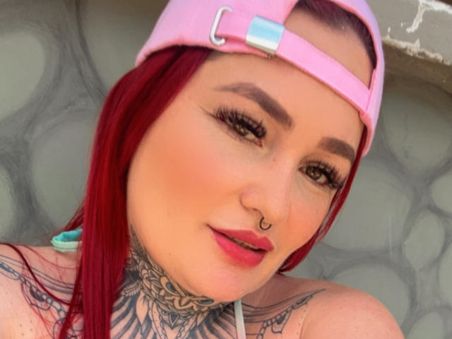 IsabellaDankworth with red hair, pink cap, nose ring, and tattoos, showing off her tits with a confident smile. Her makeup includes bold eyeliner and pink lipstick.