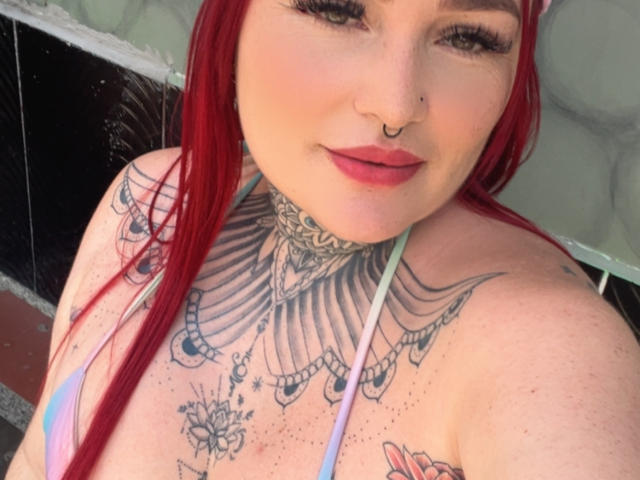 Isabella Dankworth ha i capelli rossi, un piercing al naso e un grande tatuaggio sul petto con un disegno a ventaglio. Indossa un top da bikini rosa e verde. Il suo seno è visibile..