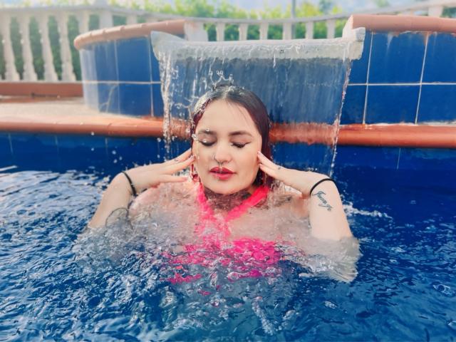 Isabella Dankworth, con i seni parzialmente scoperti, emerge dalla piscina azzurra, con l'acqua che le cola dai capelli, le mani sul viso, indossa una maglietta rosa e si vedono dei tatuaggi, con gli occhi chiusi..