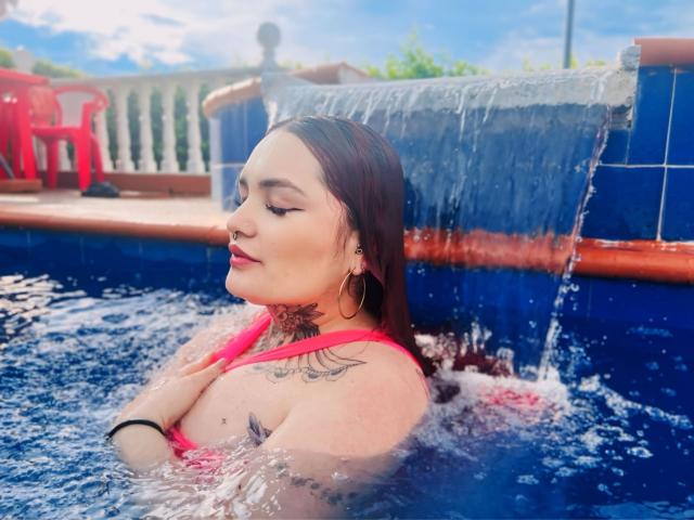 Isabella Dankworth, con i capelli castani bagnati, un top da bikini rosa e tatuaggi, seduta in una piscina blu, con l'acqua che scorre da una parete piastrellata, occhi chiusi, labbra rosa, orecchini a cerchio..