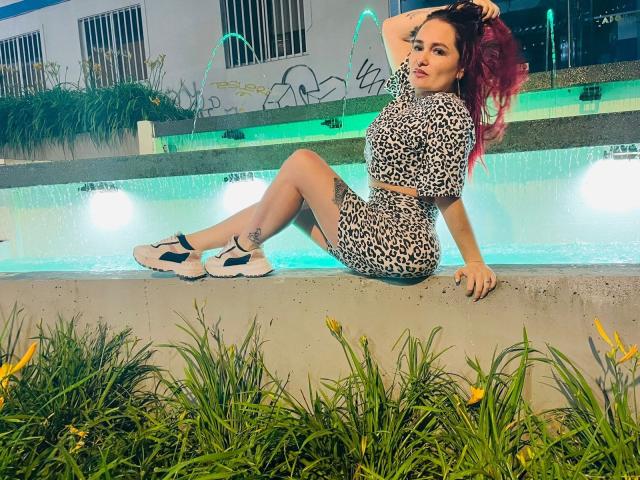 Isabella Dankworth, con i capelli rosa, un top corto e pantaloncini a stampa leopardo, seduta accanto a una piscina illuminata, con le gambe piegate, scarpe bianche e blu, una mano tra i capelli, una pianta verde in primo piano..