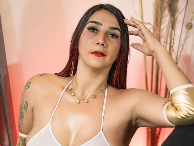 GabrielaMilf avec des cheveux roux et noirs, du rouge à lèvres rouge, un haut blanc à encolure dégagée, un bracelet en or, un collier, des tatouages, une peau brillante, une main levée, une expression confiante, des seins de taille moyenne, une lumière chaude, un flou doux..