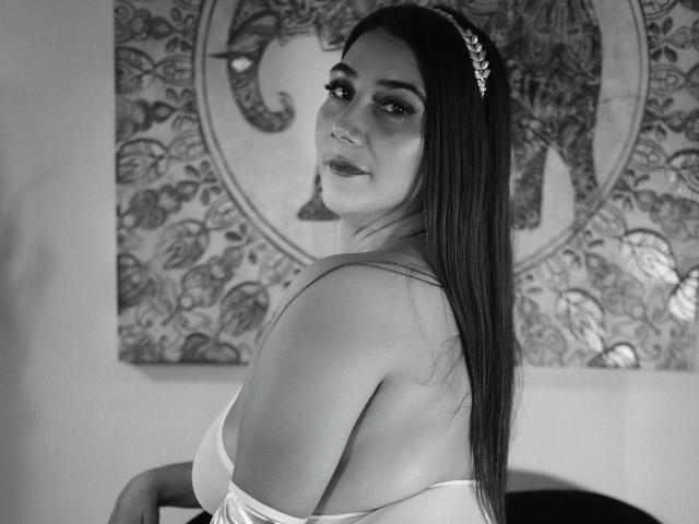 GabrielaMilf with long dark hair, wearing a white off-shoulder dress, looking over her shoulder, soft smile, decorative headpiece, black and white photo, showing her tits slightly.