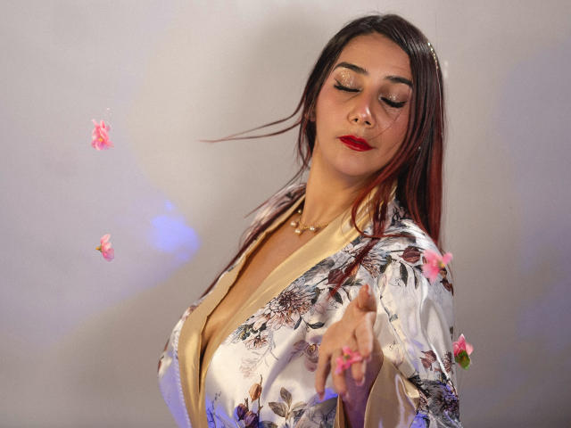GabrielaMilf with red lipstick, floral silk robe, and gold necklace, eyes closed, pointing forward, pink flowers floating, long brown hair with red tips, confident expression.