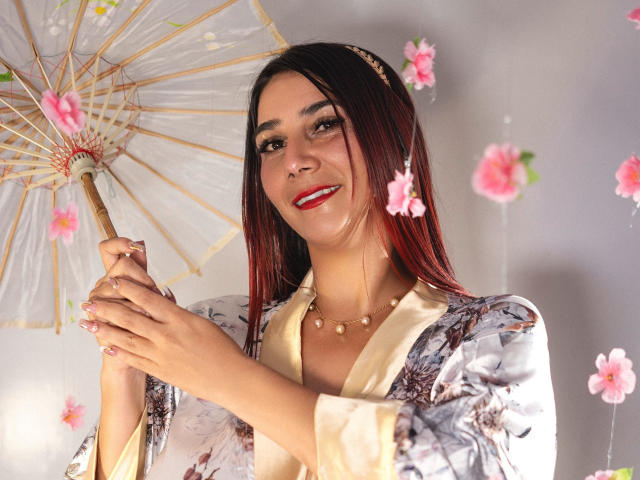 GabrielaMilf, avec des cheveux noirs aux pointes rouges, souriante, tenant un parasol japonais fleuri, portant un kimono fleuri, un collier en or et du rouge à lèvres, avec des fleurs roses autour d'elle..