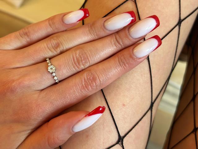 La main de Karish, avec son vernis à ongles rouge et blanc, portant une bague en diamant, repose sur sa jambe, où l'on peut voir des bas résille noirs. Ses ongles sont longs et brillants..