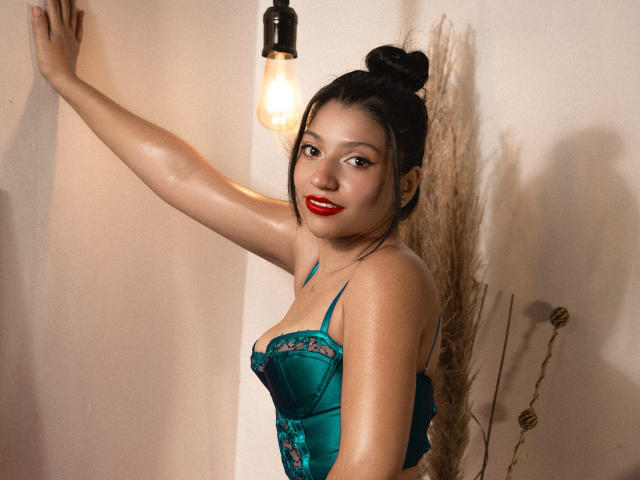 GagPrinceess with shiny skin, red lipstick, and black hair in a bun, wearing a teal lace-up lingerie, arm raised, standing next to a light bulb, smiling.