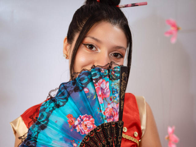 GagPrinceess holding a colorful fan with floral patterns, partially covering her face, wearing a red and beige dress, black lace trim, and a hairpin, with pink butterflies in background.