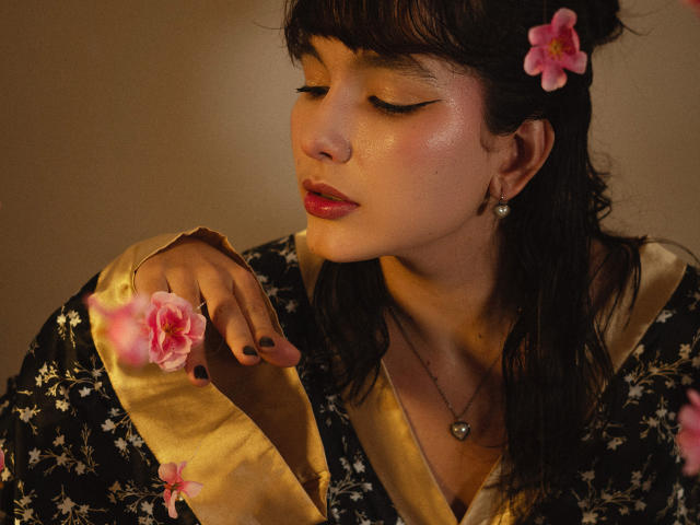 HairyValeri with pink flower in hair, eyes closed, wearing black floral robe with gold trim, pink flower on sleeve, dark nail polish, pearl earrings, and red lipstick.