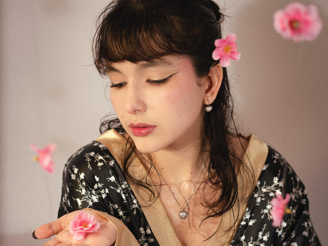 HairyValeri with closed eyes, holding pink flower, wearing black floral top, pearl earrings, heart necklace, dark hair, pink eyeliner, soft pink lips, pink flowers in background.