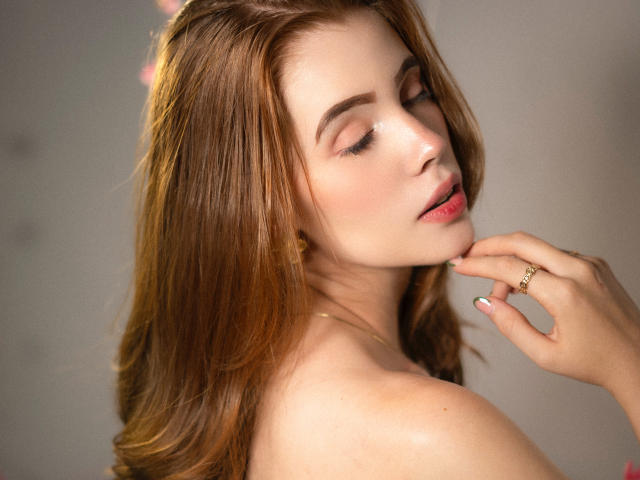 ArielLoveX with long brown hair, closed eyes, shiny skin, and pink lips, touching her chin with a gold ring, showing off her tits. Soft lighting.