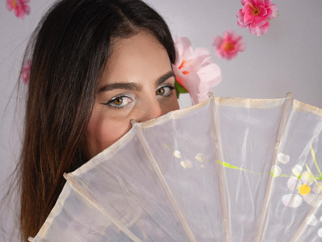 Los ojos marrones de SexxyNikole asoman detrás de un abanico delicado y semitransparente con motivos florales, su cabello oscuro cae sobre sus hombros, y hay flores rosas en el fondo..