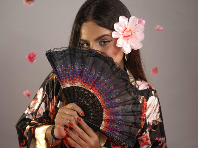 SexxyNikole with long dark hair, pink flower in hair, holding black fan with colorful edges, wearing floral kimono, red nail polish, gray background, pink flowers, and blue bracelet.