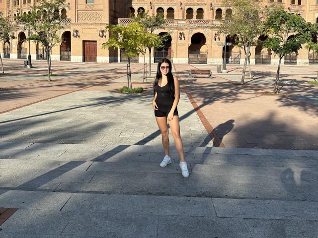 AmelieMuller stands in a sunlit plaza, wearing a black sleeveless top, black shorts, white sneakers, and sunglasses, with a hand on her hip, shadow cast on the ground.