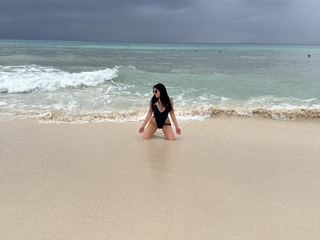 AmelieMuller kneels in shallow beach water, wearing a black one-piece, her long dark hair wet, ocean waves behind her, overcast sky, sand reflecting her silhouette.