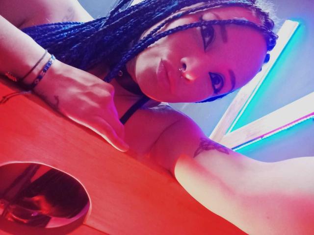 Dalila Musa mit blauen Zöpfen, Nasenring und schwarzem Choker, die eine Gitarre hält und ein Tattoo unter dem Arm zeigt, in farbenfrohem Neonlicht. Die Brüste sind nicht sichtbar. Sie lächelt in die Kamera..