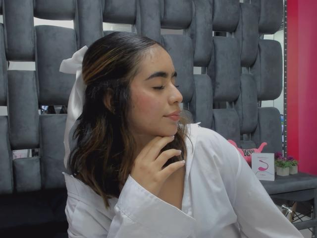 OlivaStellas with dark brown hair, white bow, and white shirt, gently touching her chin, eyes closed, blushing, sitting in front of grey, cushioned chairs.