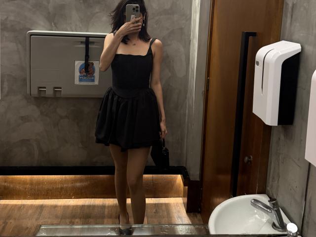 Davinaa in a black dress with a strapless top and flared skirt, standing in a bathroom, holding a phone, mirror selfie, short dark hair, tan skin, and high heels.