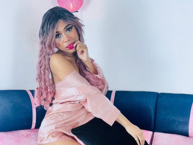 SalomeBenz with pink ombre hair, red lipstick, and pink velvet dress, sitting on blue and pink sofa, holding black pillow, looking at camera with finger near mouth.