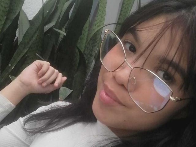 MidnightHarpy with black hair, glasses, and nose ring, smiling slightly, holding up a hand, wearing a white shirt, in a selfie. Her ass is not visible.