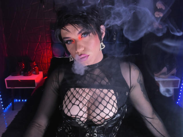 BellaFetishist with short black hair, blue eyes, and purple lipstick, smoking, wearing a black fishnet top, exposing her tits, with a black corset and earrings, in a dark room.