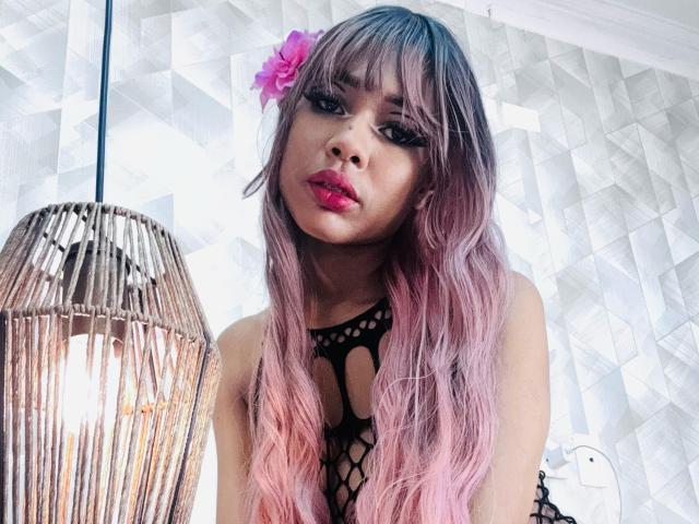 SalomeBenz with pink and purple ombre hair, black fishnet top, pink flower in hair, and red lipstick, standing next to a wicker lamp.