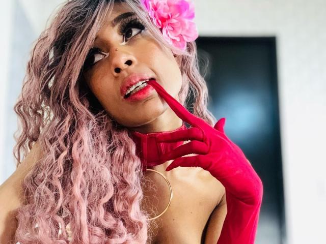 SalomeBenz with pink curly hair, red gloves, and a pink flower in her hair, biting her red-gloved finger, looking up with a seductive expression. Her tits are visible.