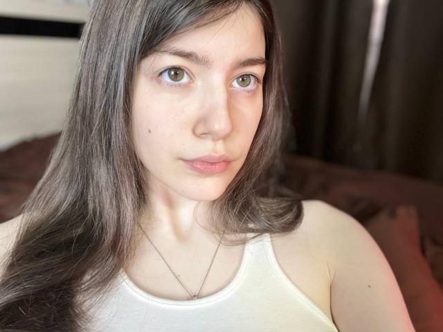 VinnaSkyHott hat hellgrüne Augen, langes braunes Haar, einen kleinen Schönheitsfleck auf ihrer linken Wange und trägt ein weißes Tanktop mit einer dünnen Halskette..