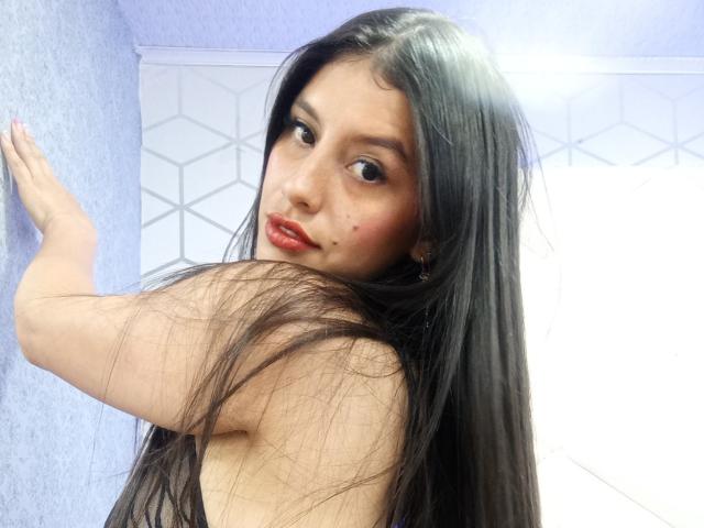 MadissonFontaine with long black hair, red lipstick, and freckles, looking back over her shoulder, touching a blue wall with her right hand, showing her bare back and shoulders.