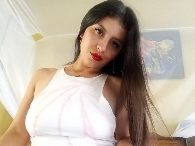MadissonFontaine with long brown hair, wearing a white sleeveless top, showing her tits, red lipstick, and a slight smile, leaning against a white wall.